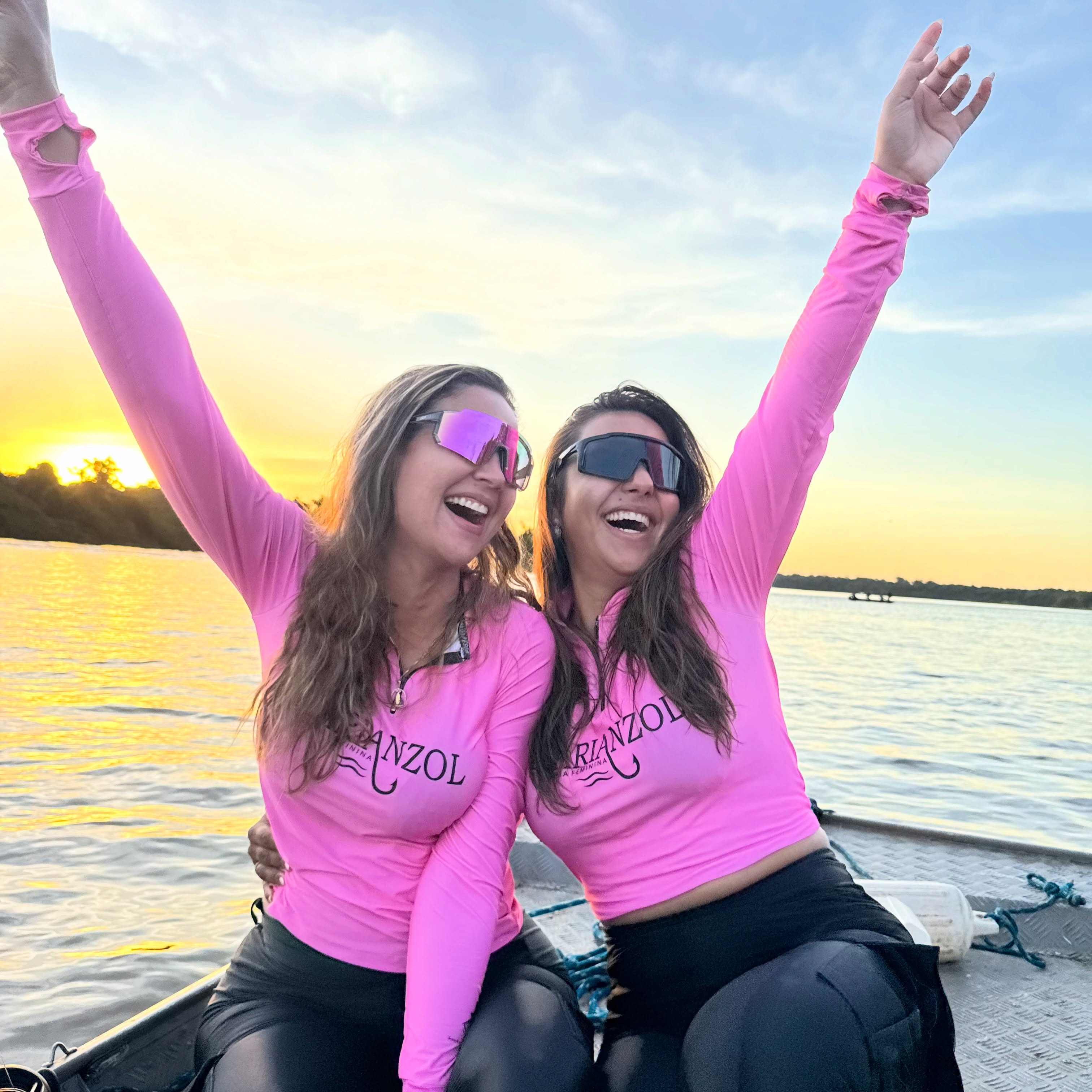 Duas mulheres com camisetas rosas da Marianzol celebrando no barco ao pôr do sol